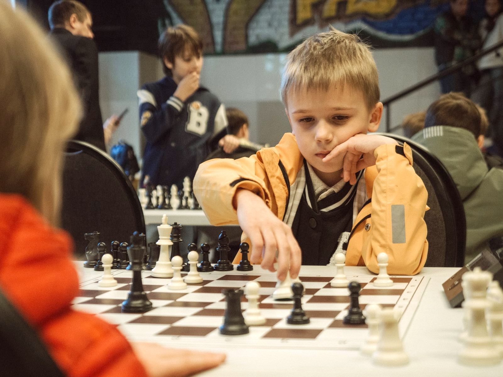Boy in a tan jacket focuses on the chessboard, reaching to move a piece during a tournament.