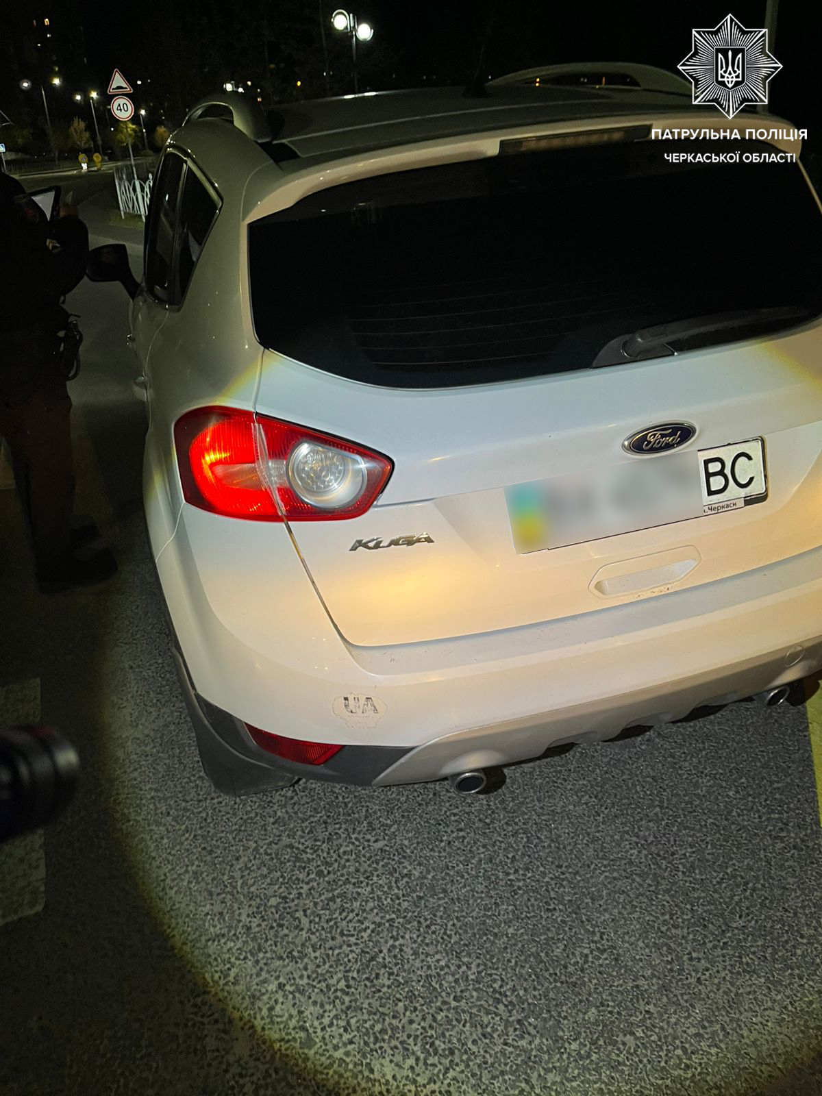 White Ford Kuga parked at night, rear view with blurred plate and police insignia watermark in top-right corner (Ukrainian patrol).