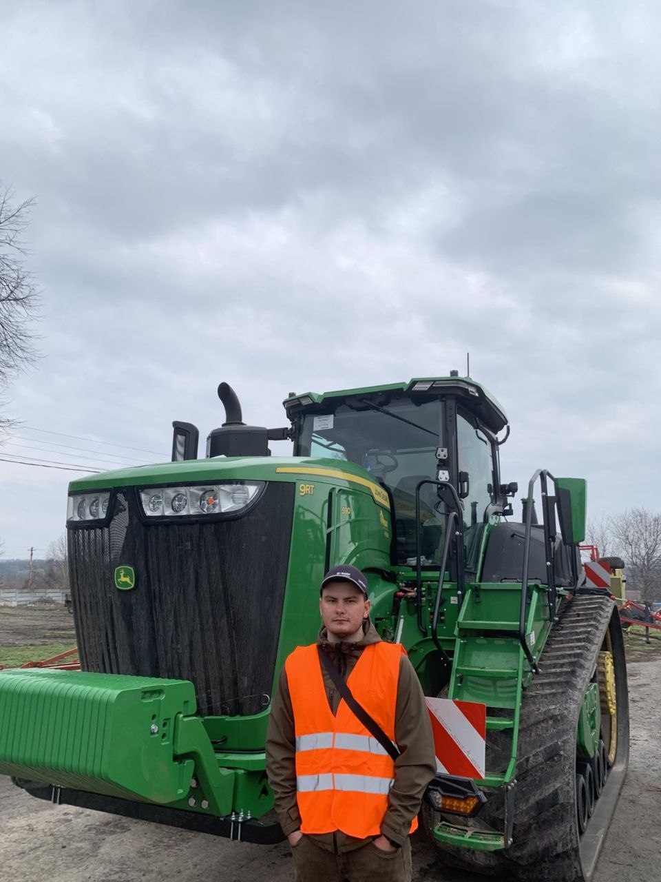 Farm worker in a bright orange safety vest stands in front of a large green John Deere combine harvester on a trailer in an overcast field.