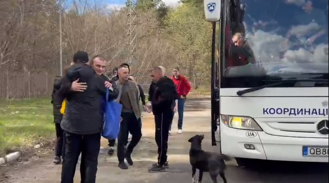 People greet and hug beside a white bus on a rural road; a black dog stands nearby as others watch in the background.