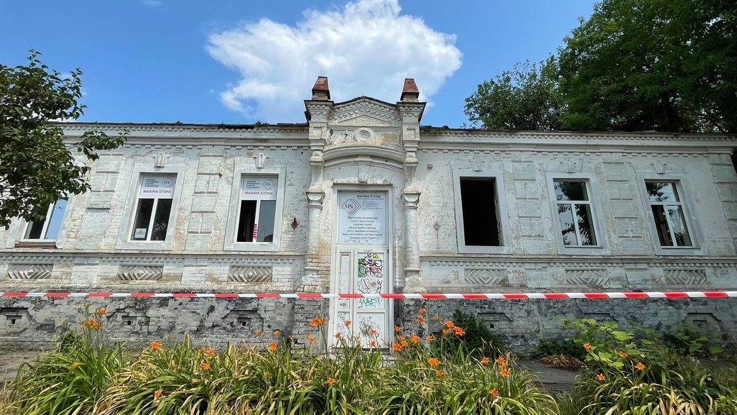 White historic building with ornate facade, red-and-white barrier tape across the front, and orange flowers in the foreground under a blue sky.