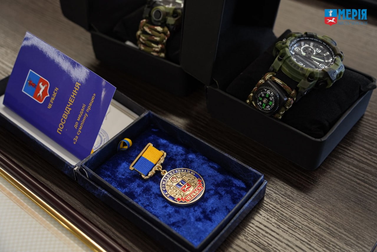 Awards display: gold medal with blue-yellow ribbon in a blue velvet box on a desk, next to a round badge and a purple certificate card. Two military-style watches sit in cases nearby.