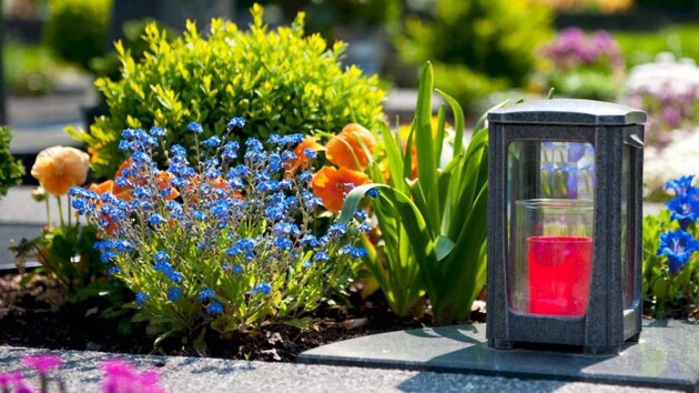 Garden bed with blue and orange flowers and a gray lantern containing a glowing red candle on a paved edge.