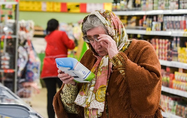 Older woman in a headscarf and brown cardigan reads a product package in a supermarket aisle, shelves visible in the background.