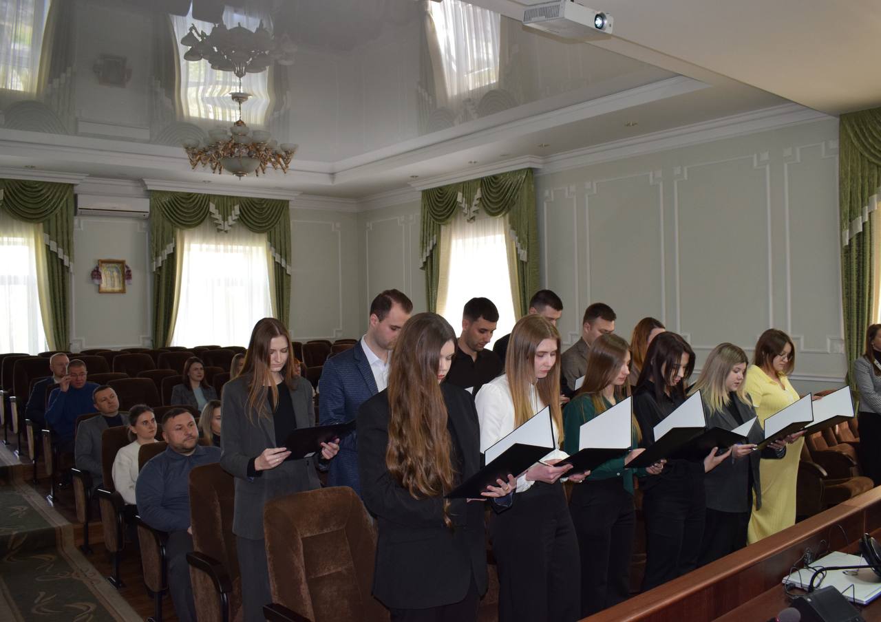 A row of young adults standing in a formal room, holding open folders as they sing or present to an audience seated in brown chairs behind them in a decorated hall.