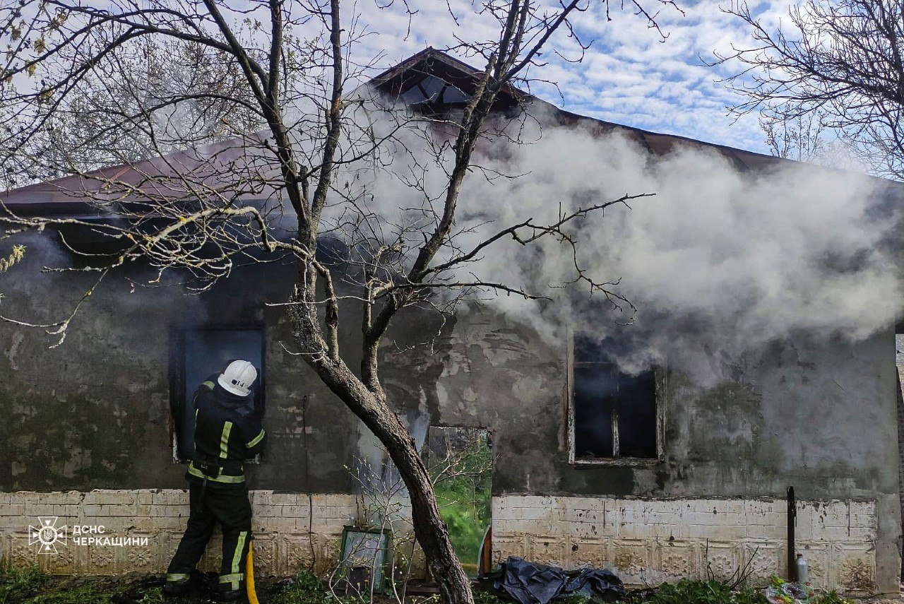 Firefighter in turnout gear uses a hose to fight a blaze in a single-story house, smoke billowing from windows and charred walls behind a leafless tree.