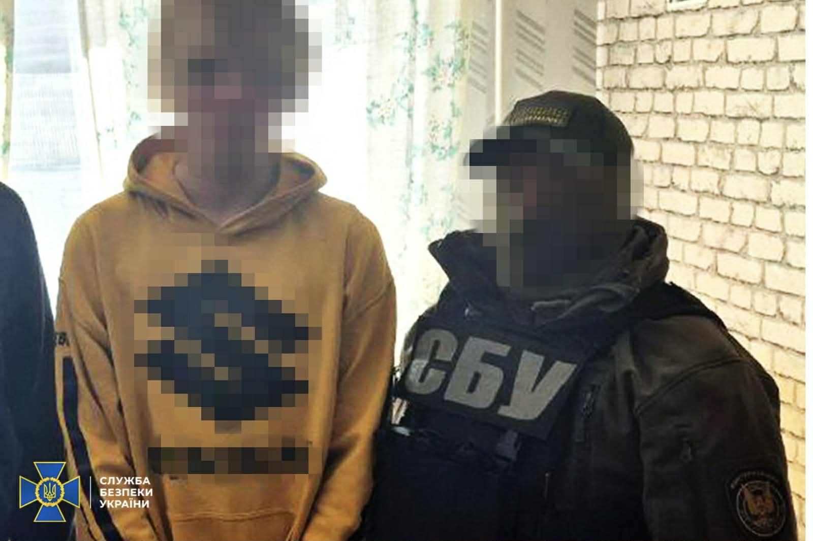 Two men indoors with faces blurred; one in a yellow hoodie with a large circular emblem, the other in a dark tactical vest marked 'СБУ' (Ukrainian Security Service).
