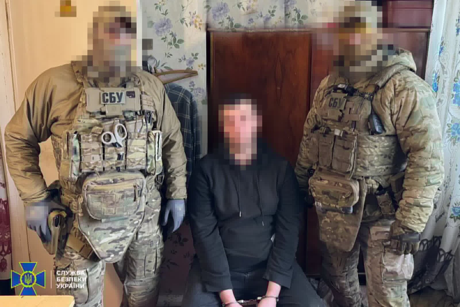 Two armed soldiers in camouflage gear flank a man in a black hoodie with his hands cuffed in front; faces pixelated; SBU Ukraine insignia bottom left.