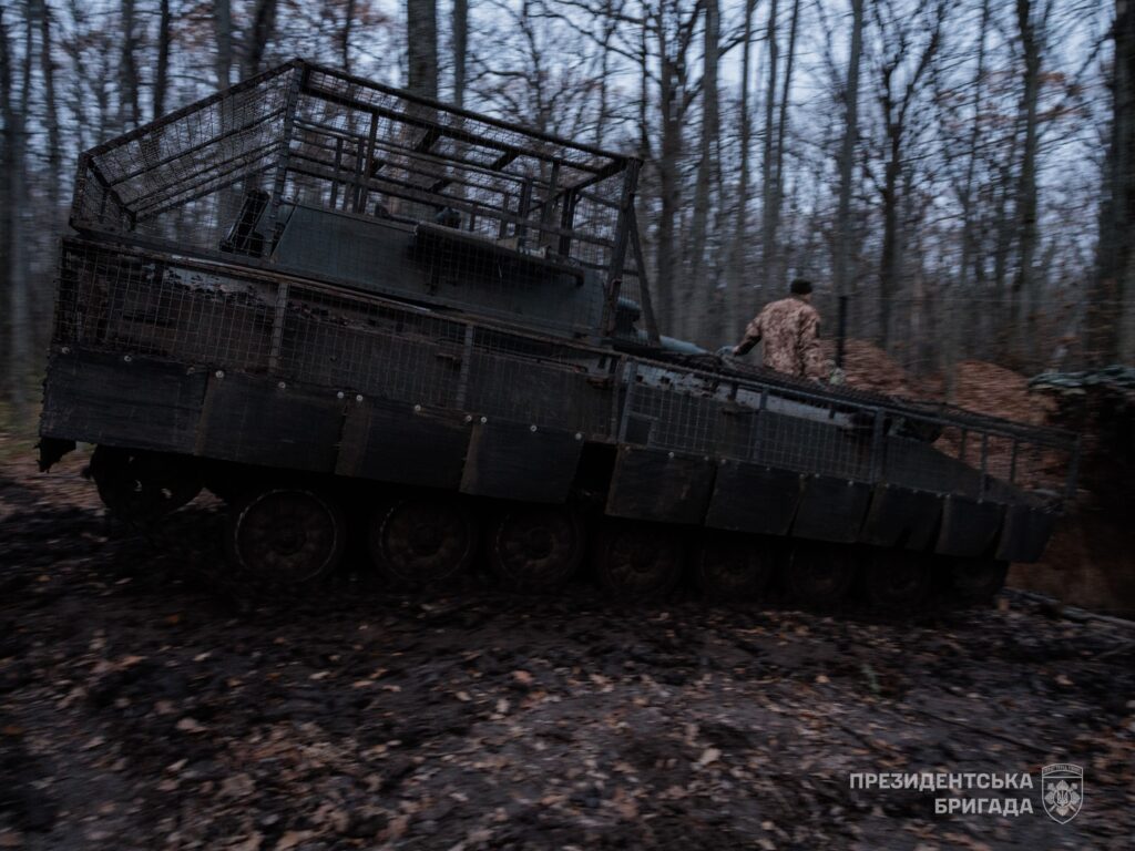 Dark armored transport on tracks in a leaf-strewn forest at dusk, with a person in camouflage standing on the side railing.