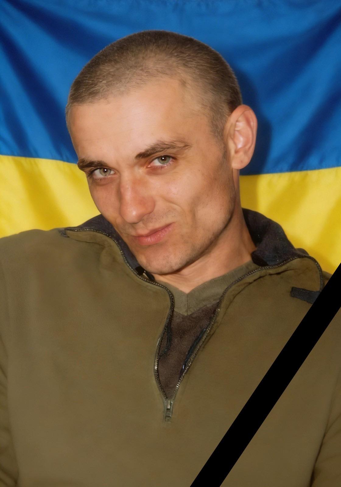 Portrait of a man with short hair in an olive jacket, in front of a blue-and-yellow Ukrainian flag with a diagonal black mourning ribbon.