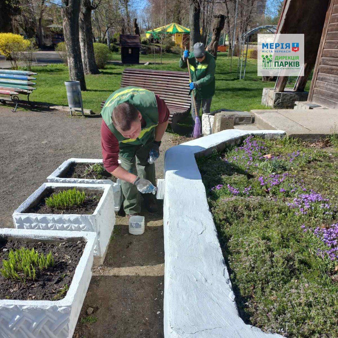 Volunteer in a green vest plants seedlings in rectangular white planters along a park path, with another person sweeping in the background and benches nearby.