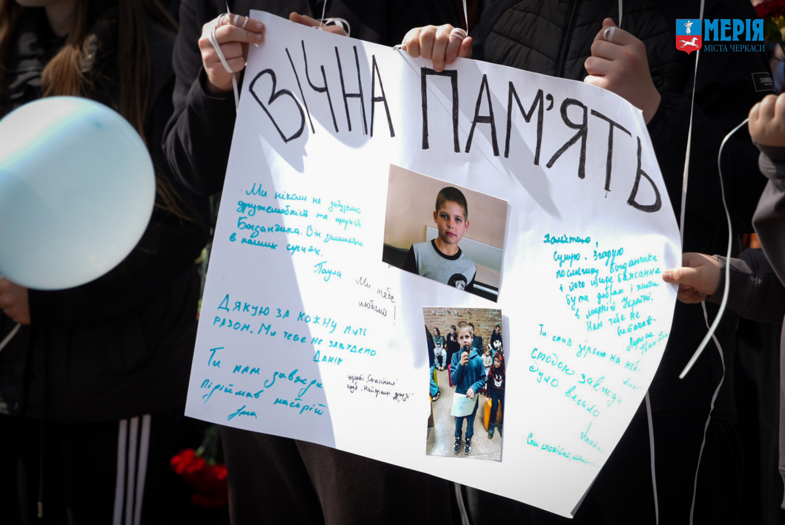 Public memorial poster reading 'Вічна памʼять' with photos of children and blue handwritten notes, held by participants at a gathering.