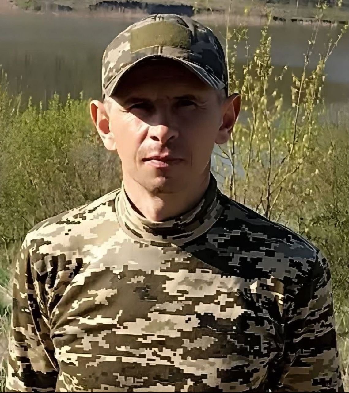 Military service member in digital camouflage uniform standing outdoors by tall grasses and a body of water in the background