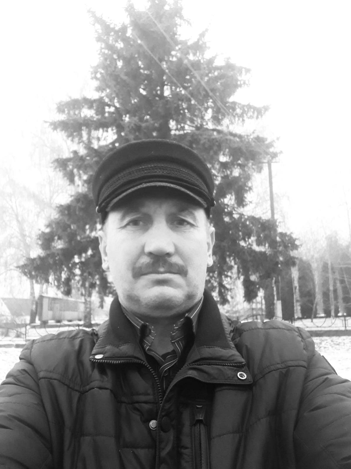 Man in a dark quilted jacket and peaked cap outdoors, facing the camera with a neutral expression, bare trees in the background.