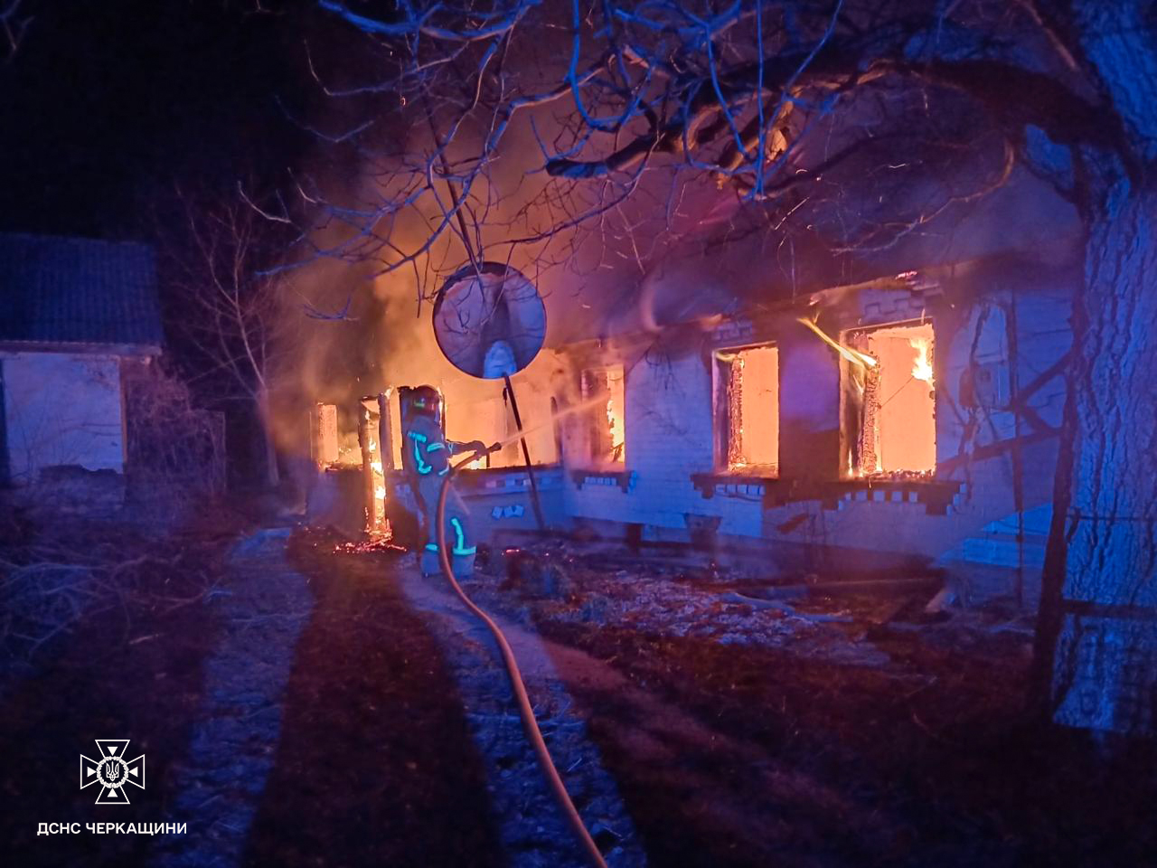 Firefighters in gear spray water on a blazing single-story house at night, windows glowing orange as flames tower from the structure.