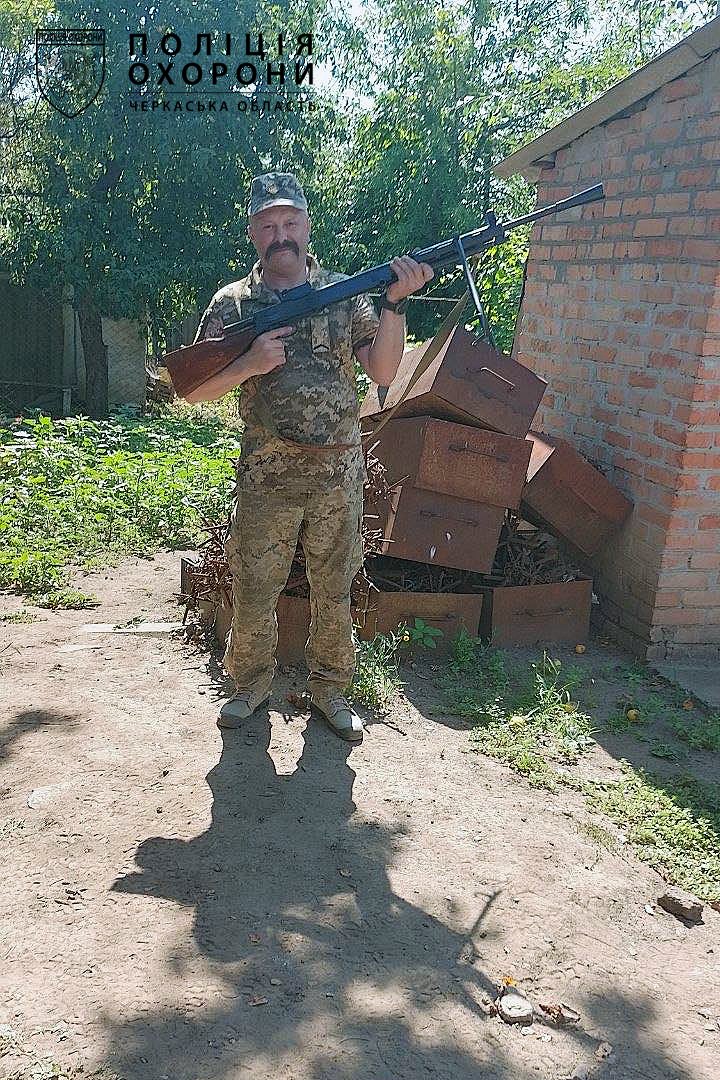 Camouflaged soldier holding a rifle outside a brick building with stacked crates nearby; logo in the top-left corner.