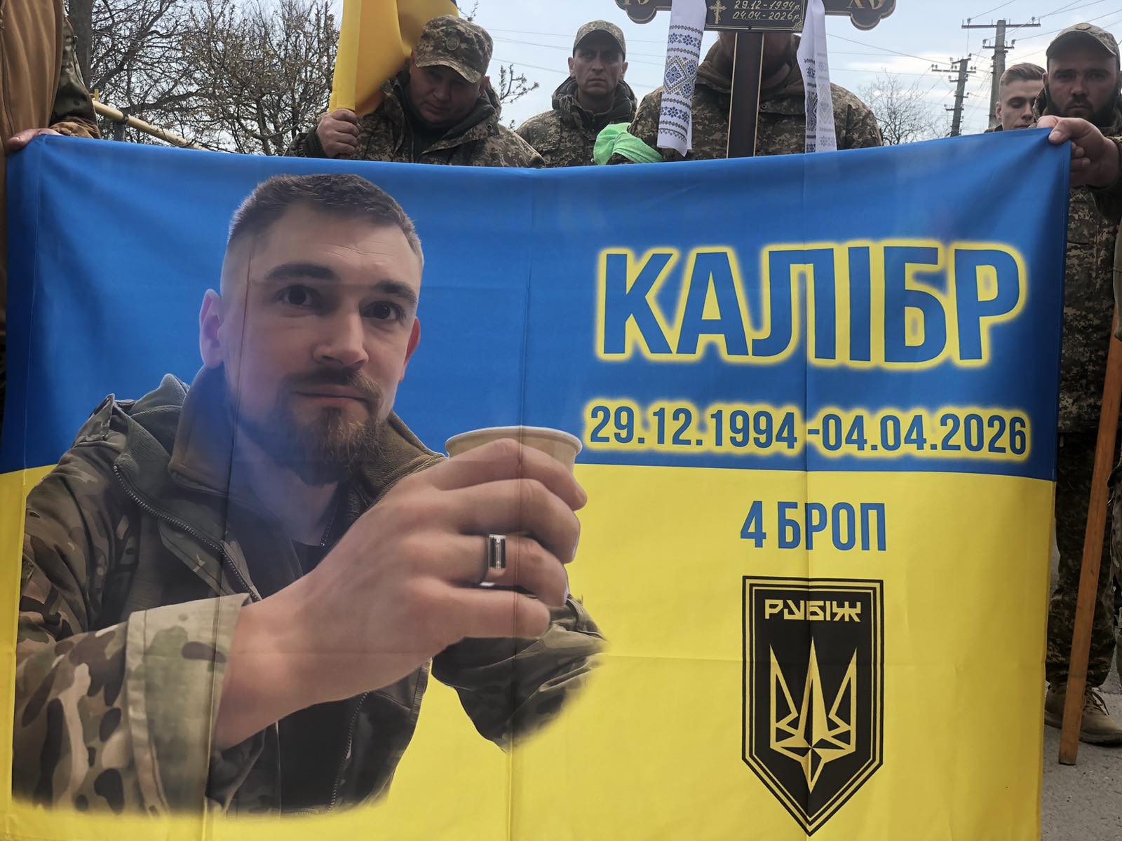 Commemorative blue-and-yellow banner held by soldiers, featuring a portrait of a man in camouflage raising a cup, with the text КАЛІБР and the dates 29.12.1994–04.04.2026, plus a shield emblem.