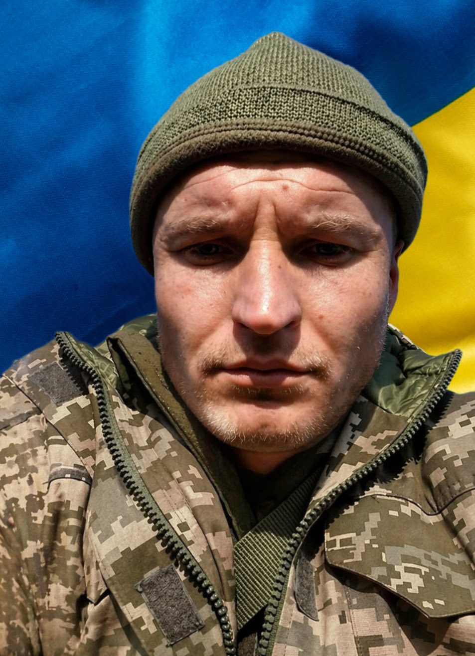 Close-up of a man in camouflage jacket and olive knit cap with a blue and yellow flag behind him.