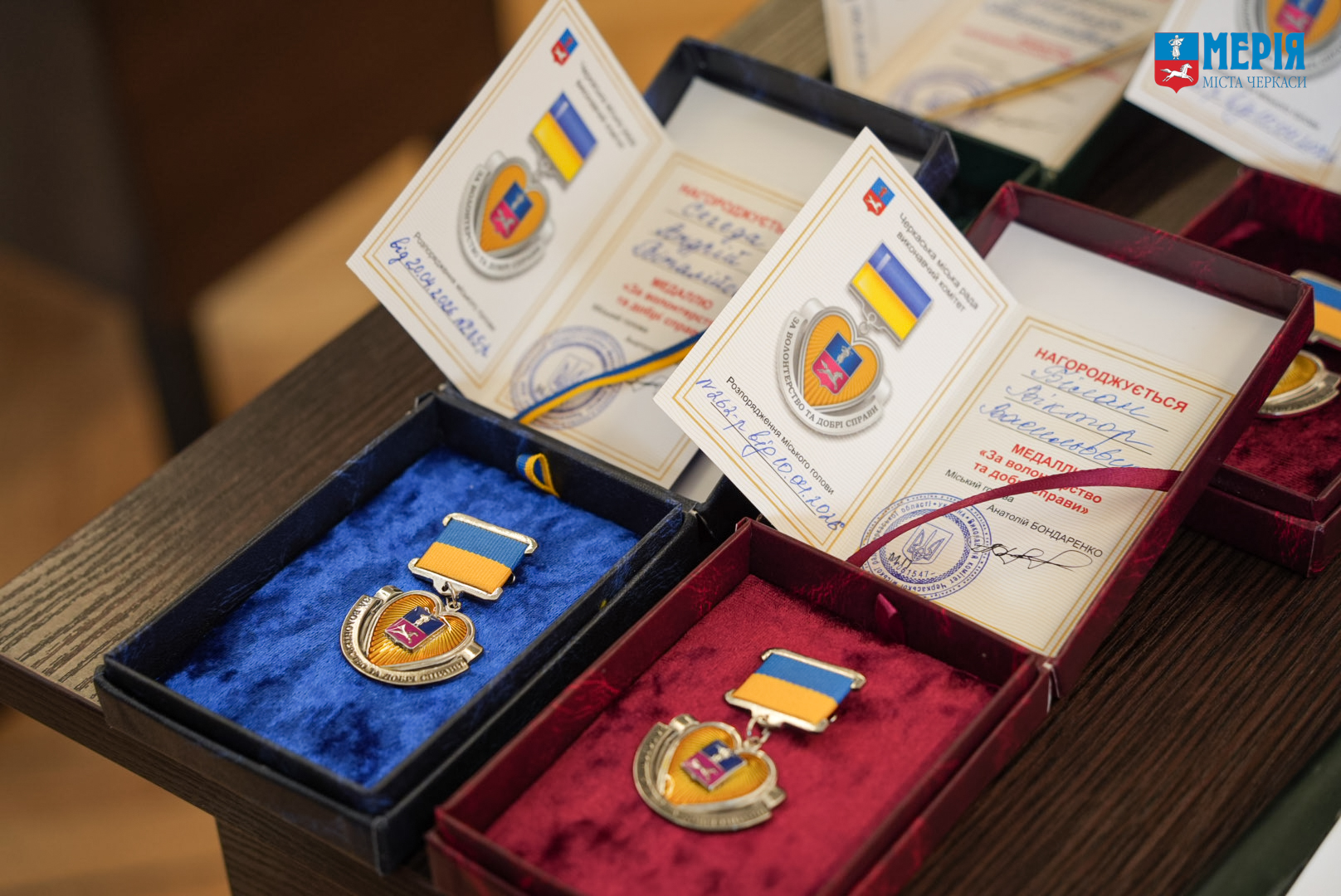 Medals with blue and yellow ribbons in blue and red velvet boxes displayed on a wooden table, alongside certificates with Ukrainian flags and seals.