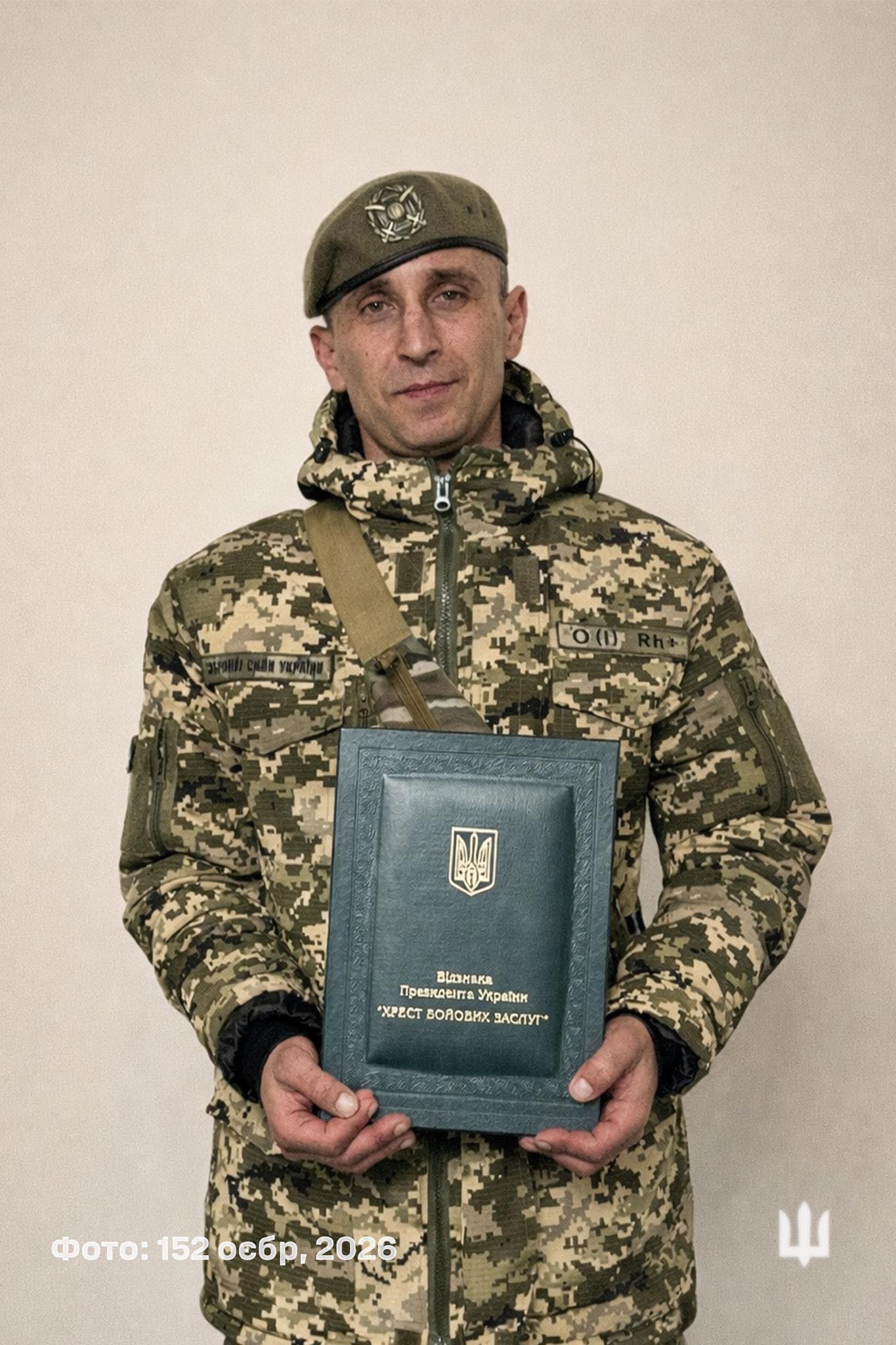 Ukrainian soldier in camouflage holding a blue book with gold coat-of-arms emblem. He wears a beret and a camouflage jacket.