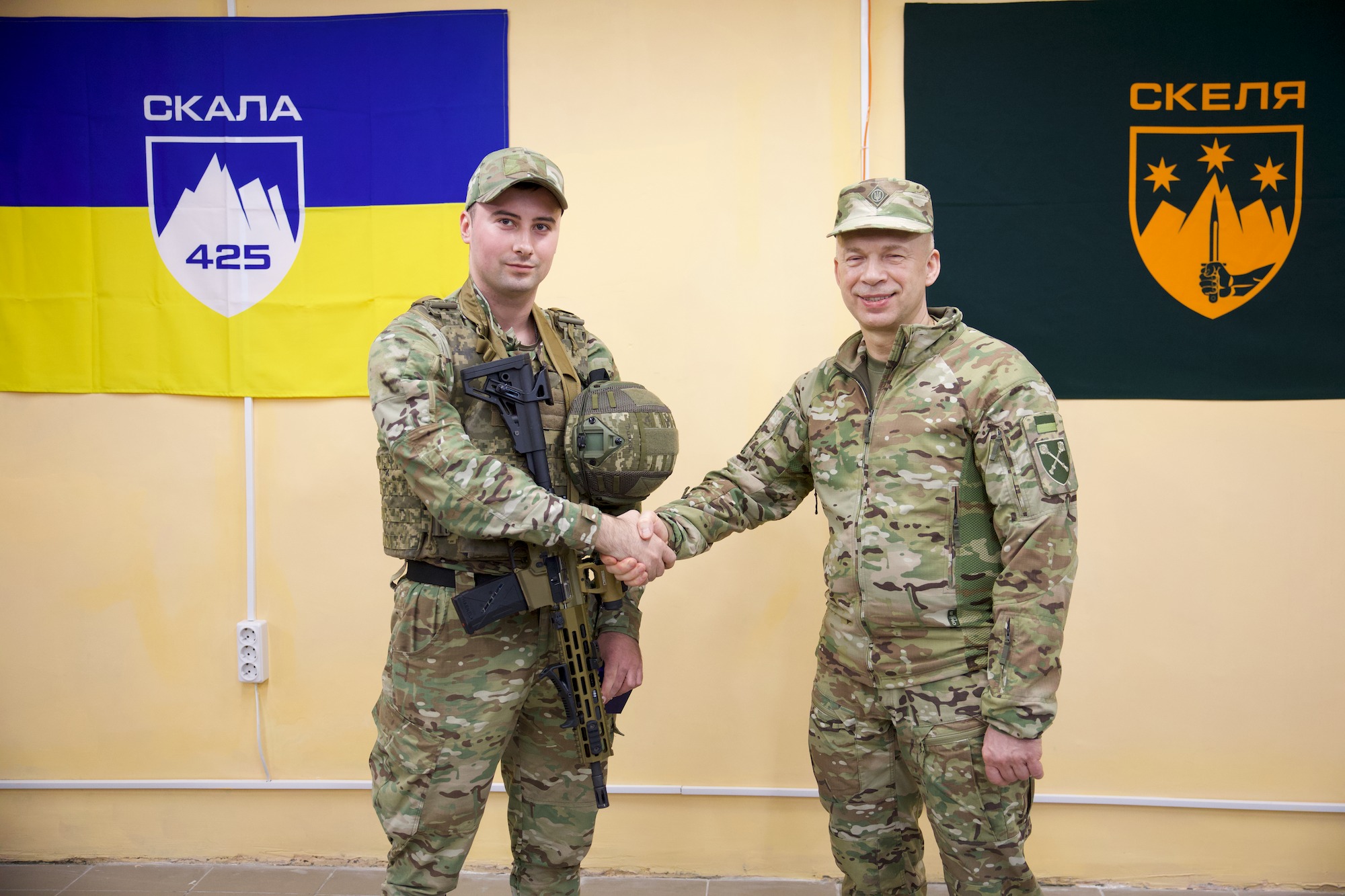 Two soldiers in camouflage shake hands in front of blue-yellow flags, symbolizing a military agreement or greeting.