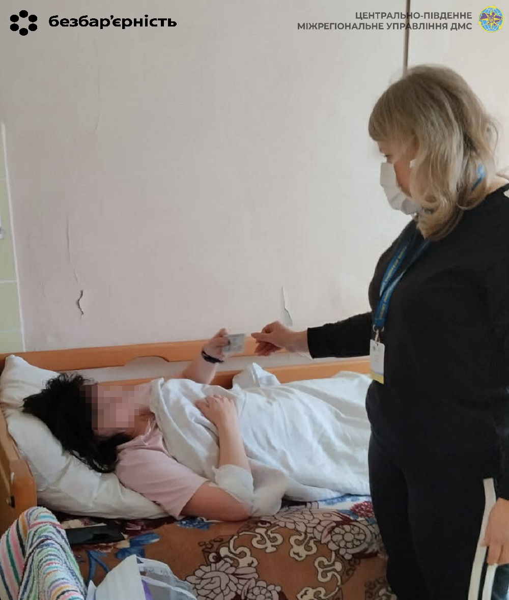 Caregiver in a mask hands a small item to a patient lying in bed in a hospital room.