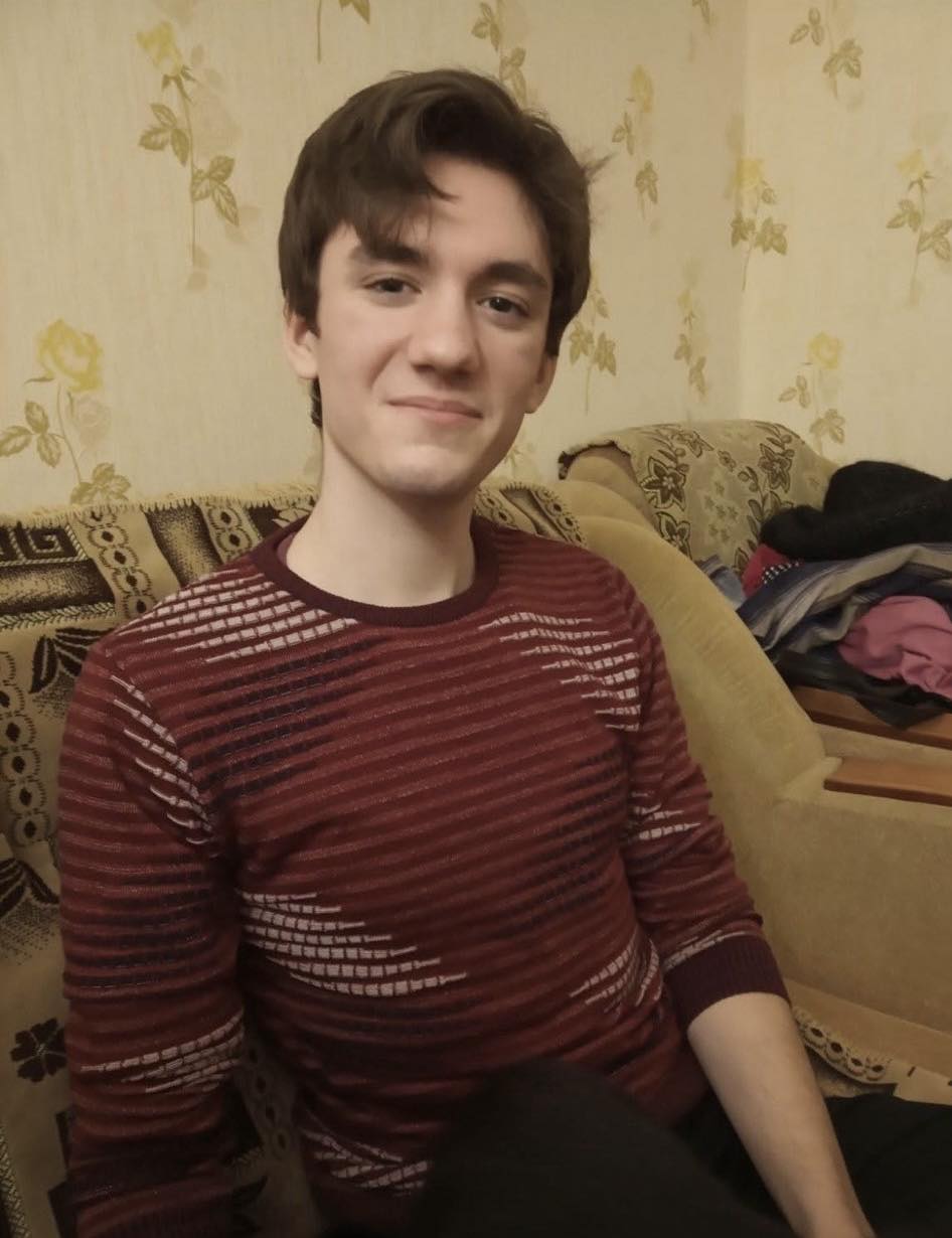 Young man sitting on a patterned sofa, smiling at the camera in a burgundy striped sweater.