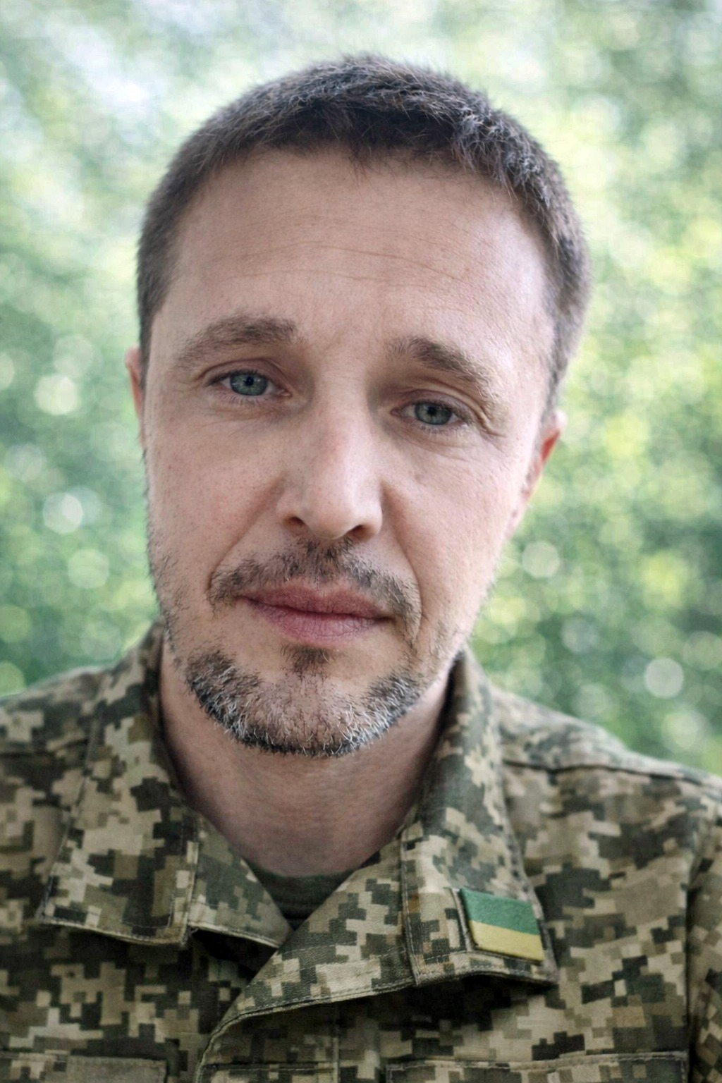 Close-up of a man in camouflage uniform with a green-yellow patch, outdoors with a leafy blurred background.