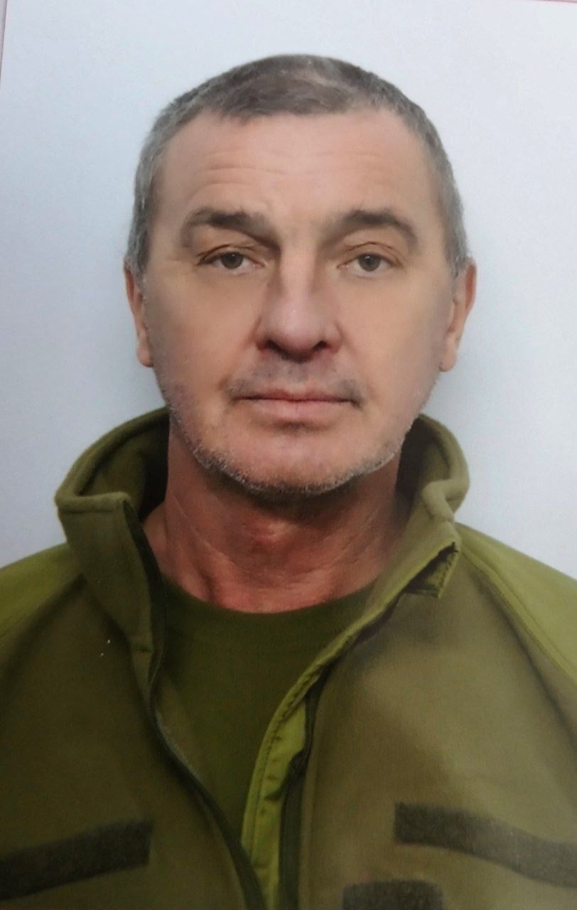 Head-and-shoulders portrait of a middle-aged man with short gray hair, wearing an olive-green jacket and a neutral expression against a light background.