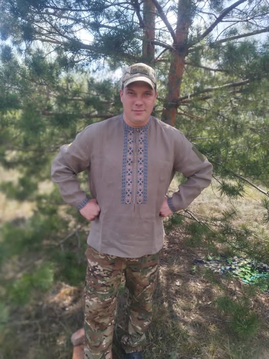 Man standing in a forest wearing a beige embroidered tunic, camouflage pants, and a cap, with hands on hips.