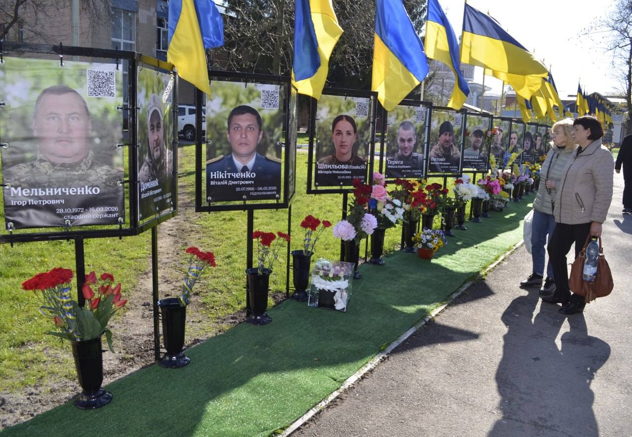 Row of framed portraits with blue-and-yellow Ukrainian flags overhead, flowers at their bases, as visitors observe a memorial display.