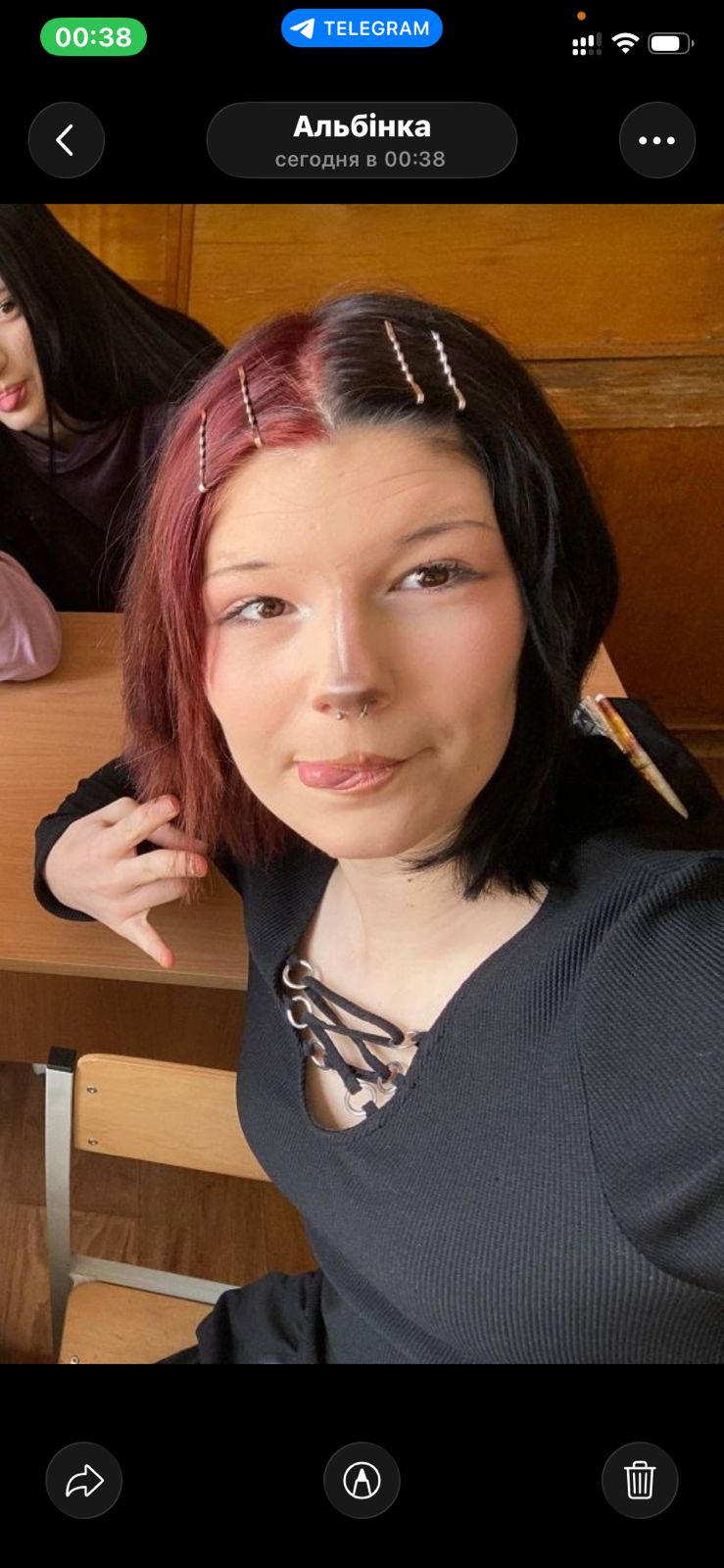 Young woman with two-tone black and red hair and hairpins, wearing a black lace-up top, sitting at a wooden desk indoors and looking to the side.
