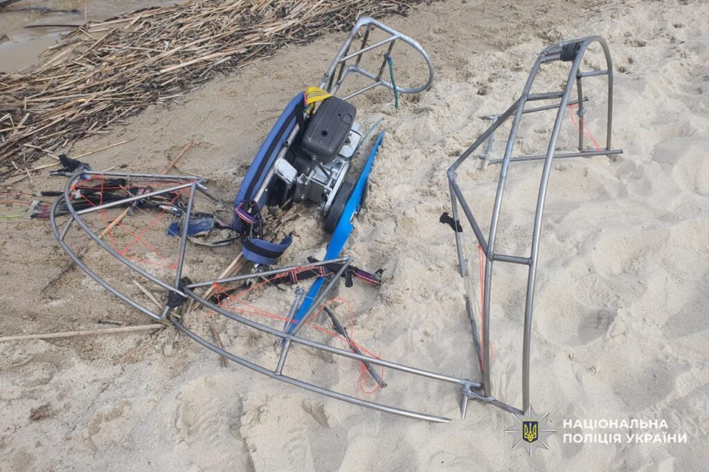Wrecked paramotor frame and engine lying on the sandy beach with tangled cords; Ukrainian police watermark in corner.