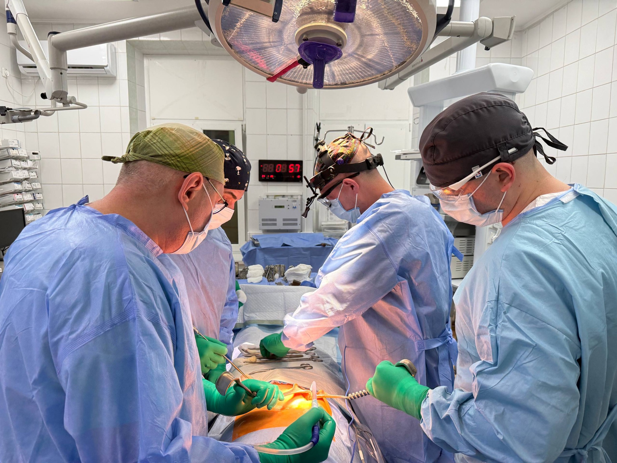 Surgeons in blue gowns and green gloves perform surgery around an operating table under bright overhead lighting in an OR.