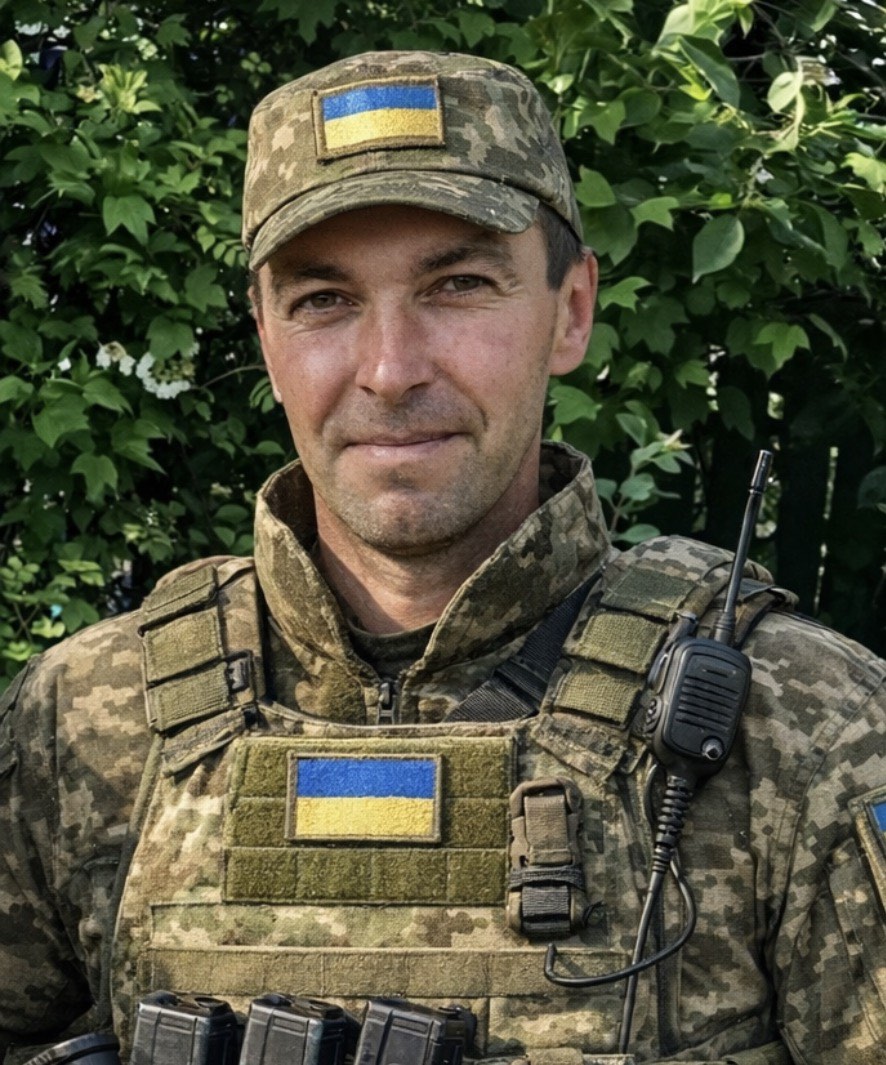Male soldier in camouflage uniform with Ukrainian flag patches, wearing tactical vest and radio, outdoors among green foliage.