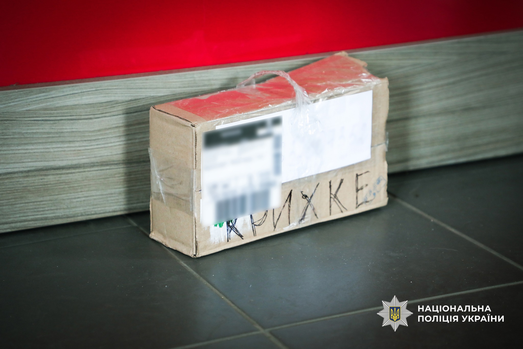 Cardboard package on a dark tiled floor, sealed with tape and plastic wrap, with blurred shipping labels and a Ukrainian police watermark in the bottom-right corner of the image.