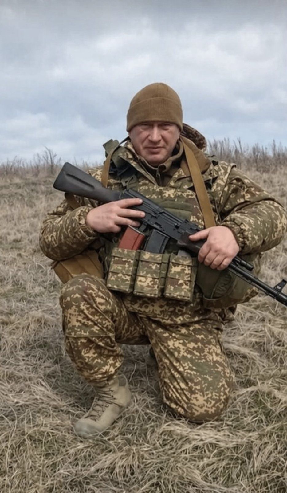 Soldier in camouflage kneeling in a field, gripping a rifle with ammunition pouches visible on his vest.