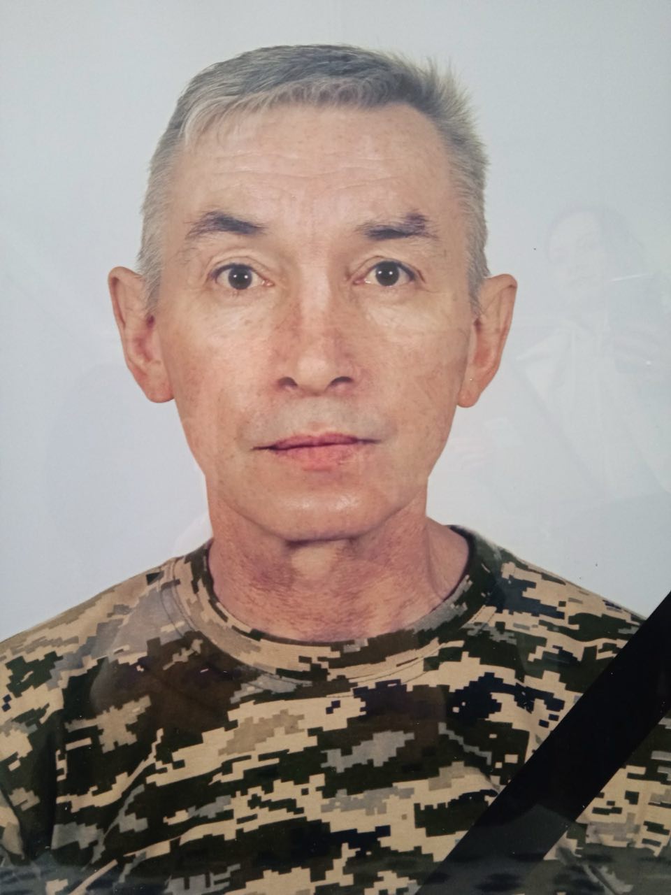 Portrait of a middle-aged man in a camouflage shirt looking straight at the camera with a neutral expression, a dark strap crossing his chest visible on the right