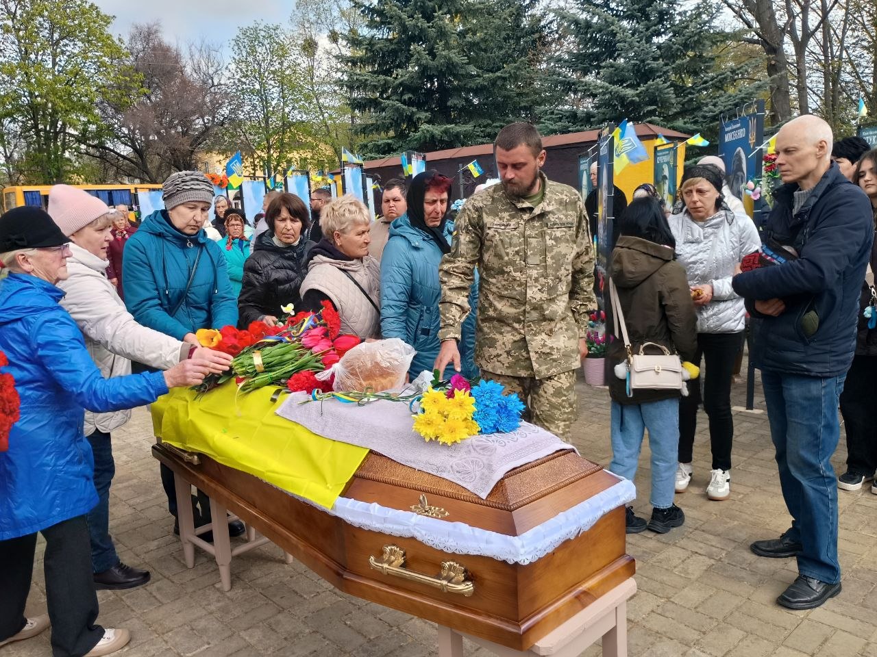 Casket on a table adorned with yellow cloth and flowers as mourners place flowers at a cemetery-like gathering with a soldier present in camouflage.