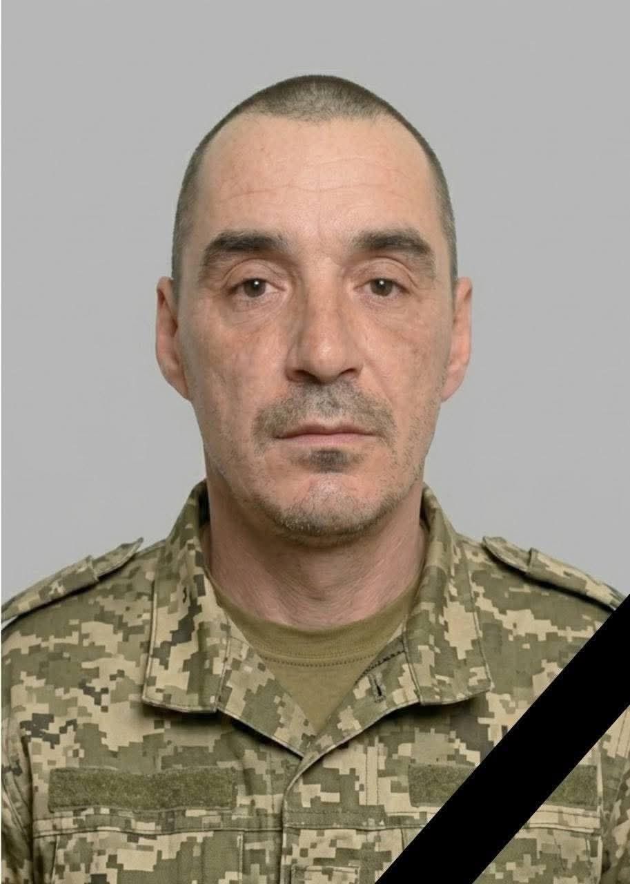 Portrait of a middle-aged man in camouflage military uniform with a black diagonal mourning ribbon across his torso, looking at the camera.