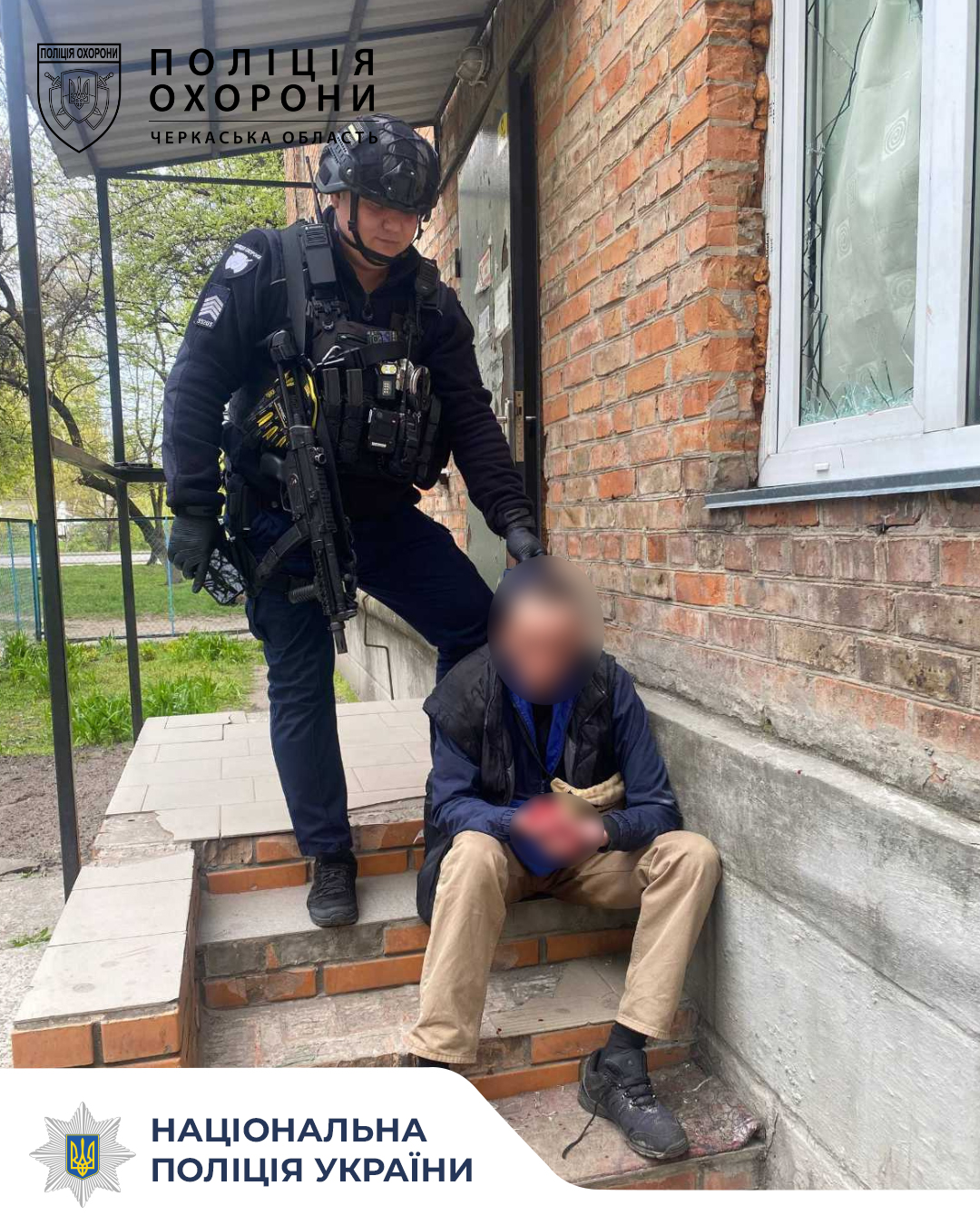 Policeman in tactical gear with a rifle standing over a seated man on brick steps outside a building; Ukrainian police insignia visible.