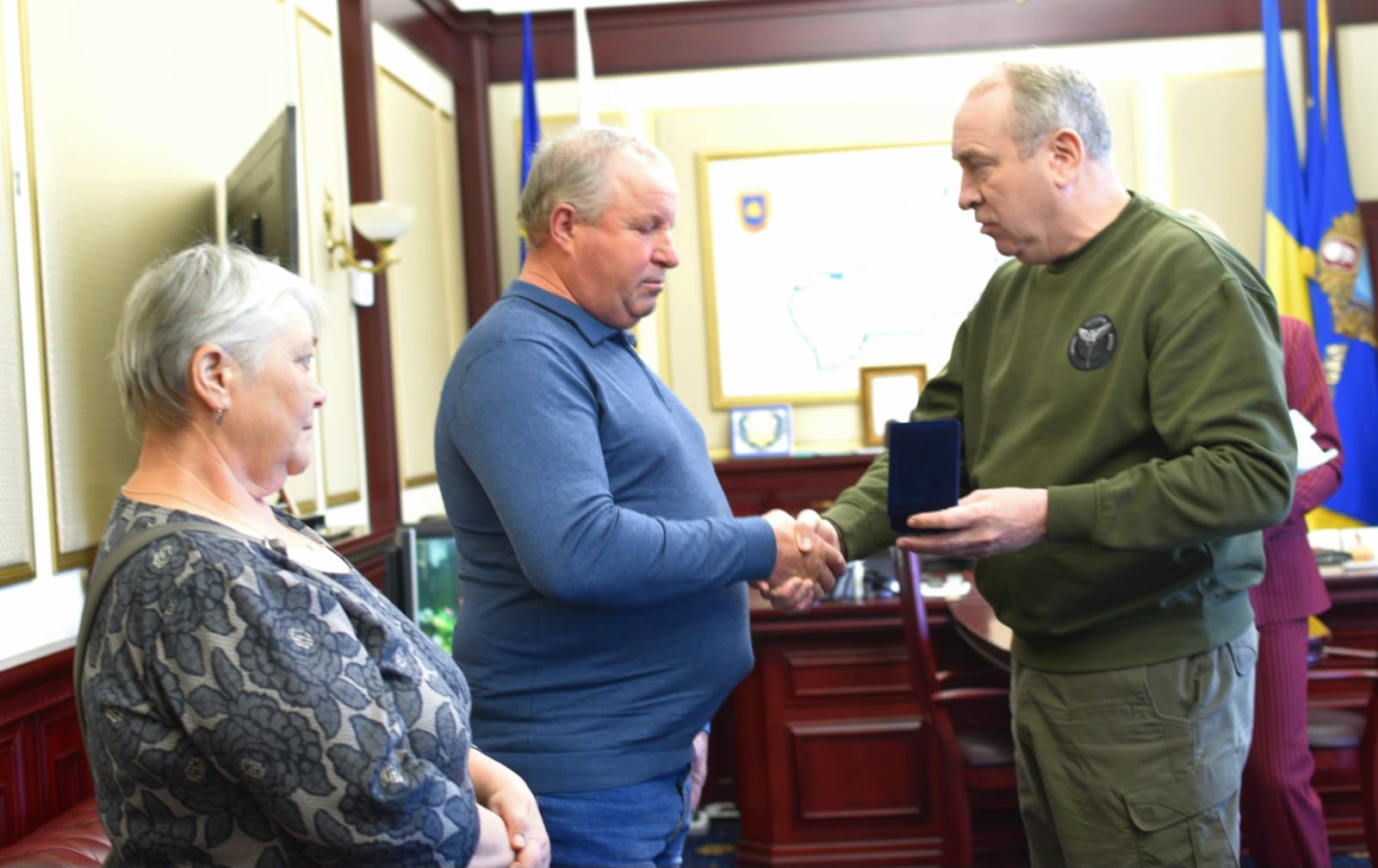 Two men shake hands in a formal award ceremony while a surprised woman sits nearby in a patterned blouse; flags and a map hang in the background.
