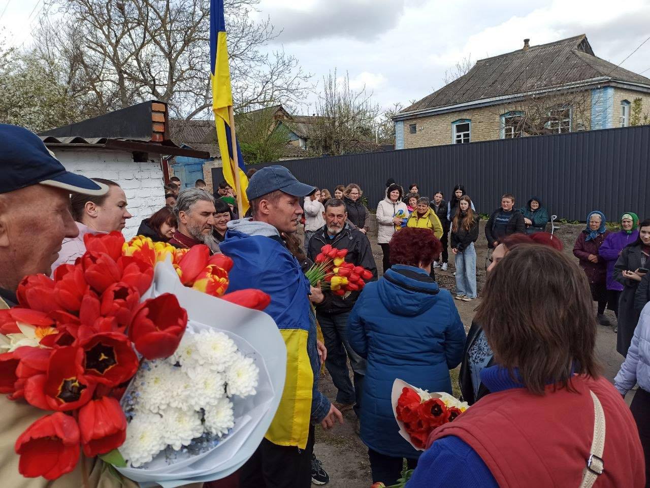 A crowd gathers in a village yard, several people holding bright red and white bouquets amid a blue-and-yellow flag backdrop.