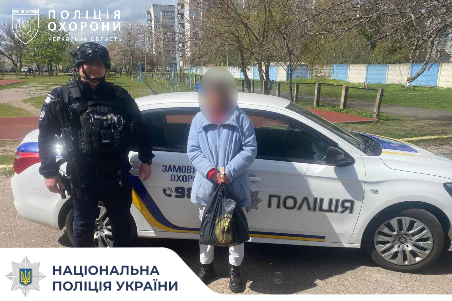 Policeman in tactical gear and helmet beside a white Ukrainian police car with blue-yellow stripes; a civilian stands in front holding a bag.