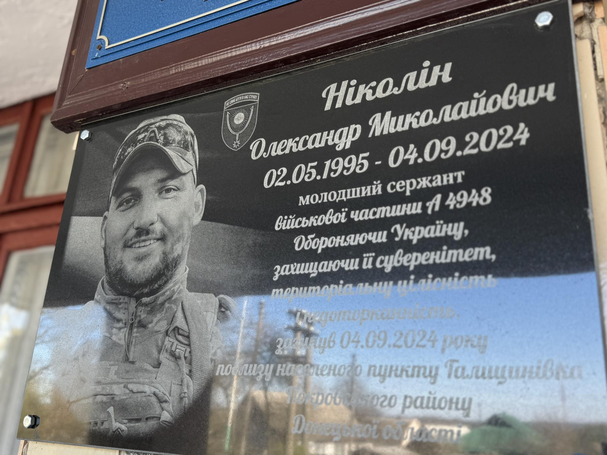 Black-and-white portrait of a smiling soldier on a commemorative plaque with Ukrainian text and emblem above the date range 1995–2024 (military memorial).