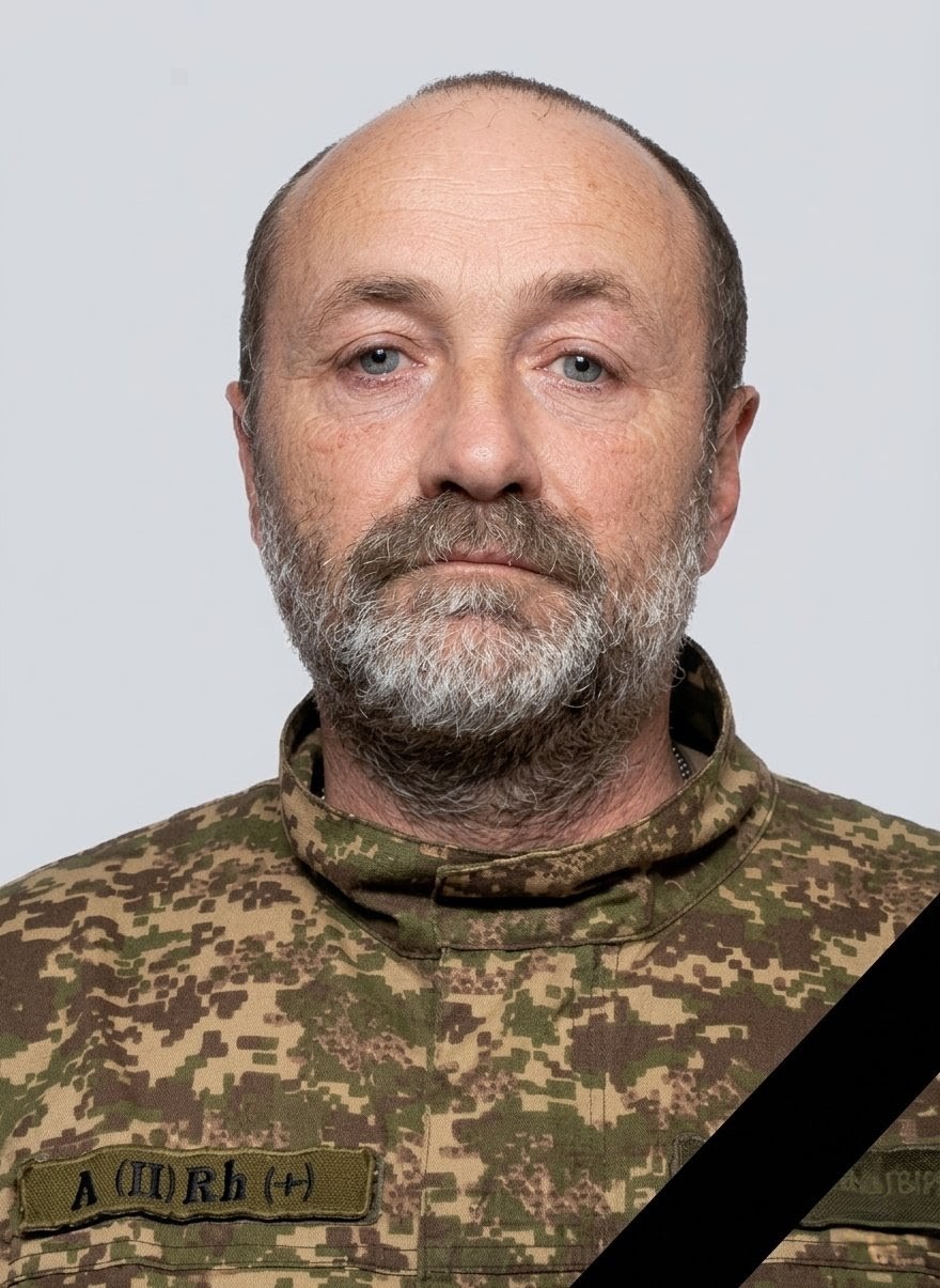 Portrait of a middle-aged man in camouflage military uniform with a black diagonal ribbon across his chest against a neutral gray background.