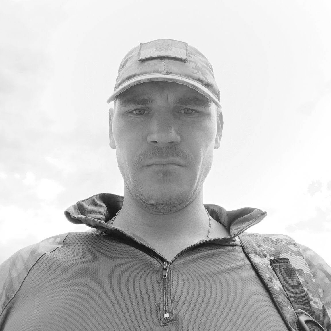 Man in camouflage cap and tactical vest looks at the camera in a chest-up outdoor portrait.