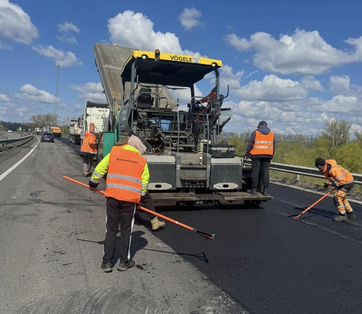 Highway construction: workers in orange vests spread fresh asphalt as a large paver lays it down, with trucks and a blue sky in the background.