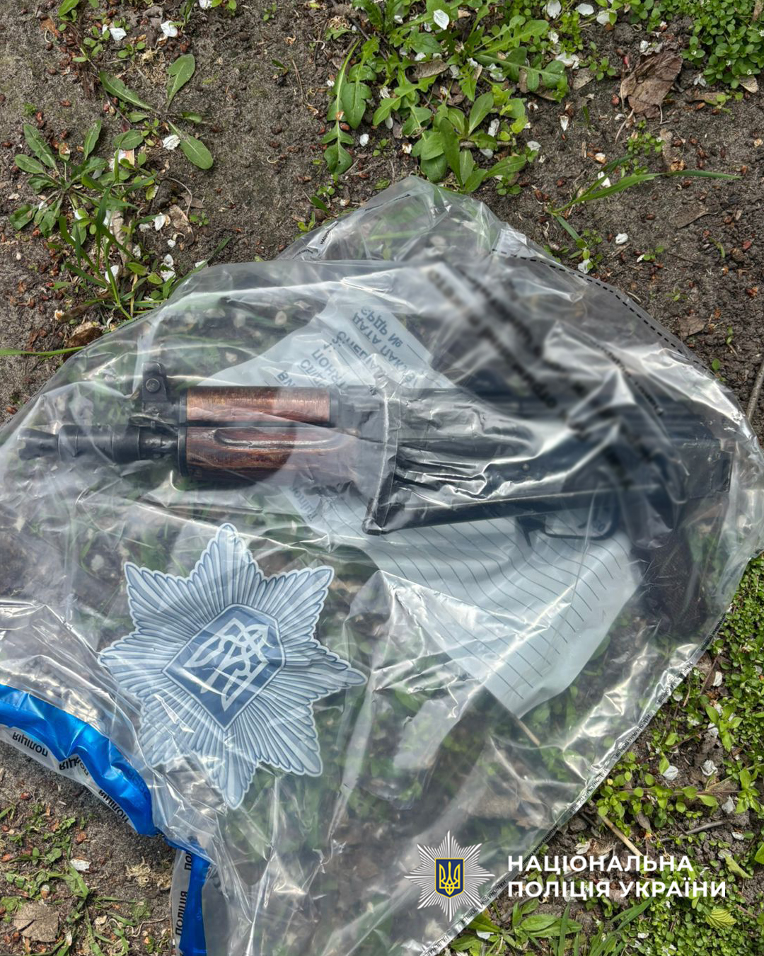 Plastic-wrapped police firearm with a wooden grip on the ground, Ukrainian police badge visible on the bag.
