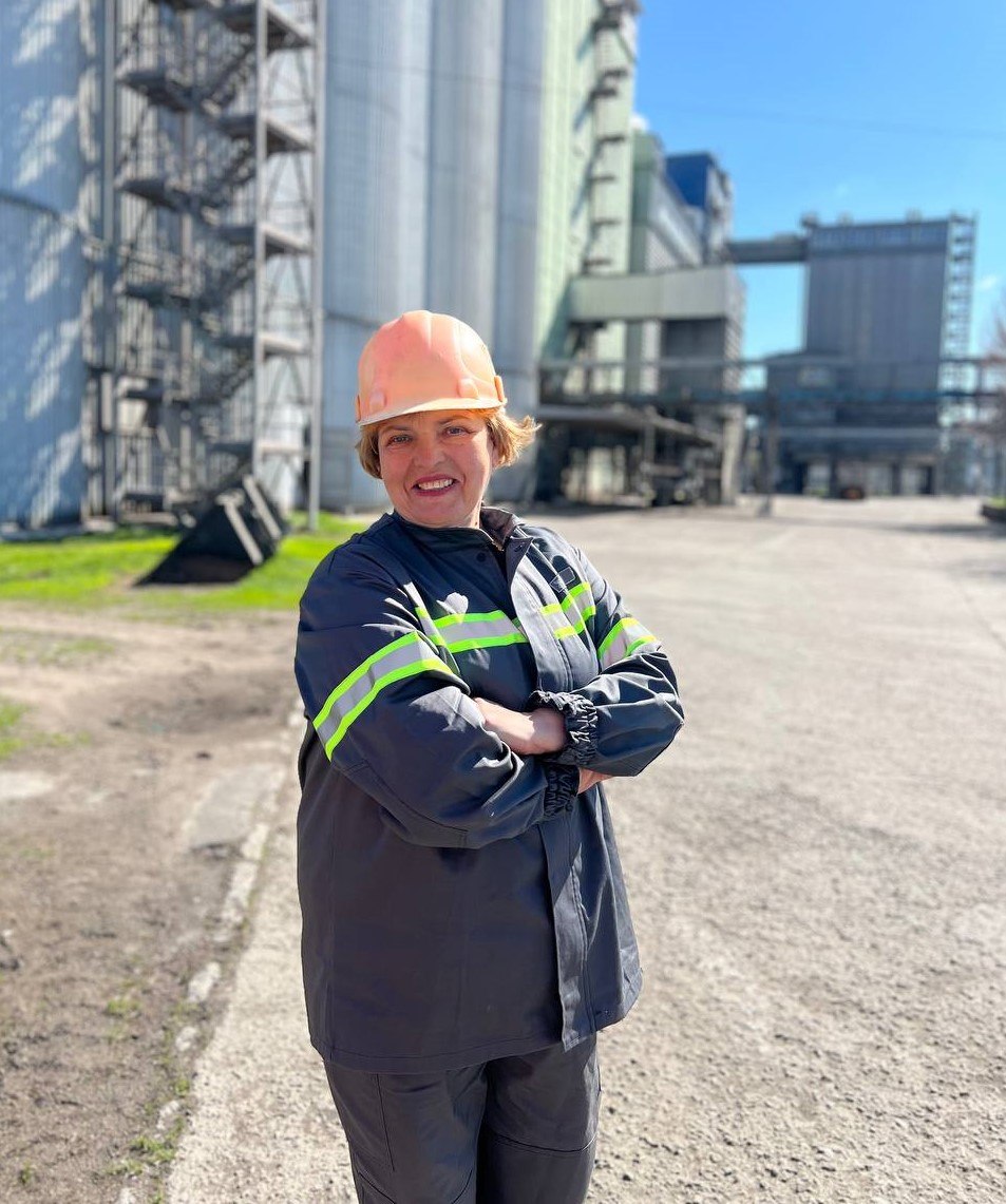Female industrial worker in an orange hard hat and dark coverall with neon safety stripes, arms crossed at a plant site.